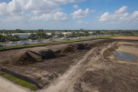 Multi-acre commercial clearing beside a wide suburban arterial in Broward, mulch windrows and silt fence with a retention pond excavation started