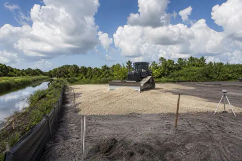 Small dozer pushing tan shellrock across a fresh Davie building pad, string lines set and erosion barrier staked at the canal side